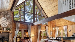 A cozy cabin interior with a stone fireplace, leather sofas, and large windows offering a view of trees. - Family Cabin Retreat: Fireplace, Game Room, Outdoor Deck, Firepit (Munds Park)