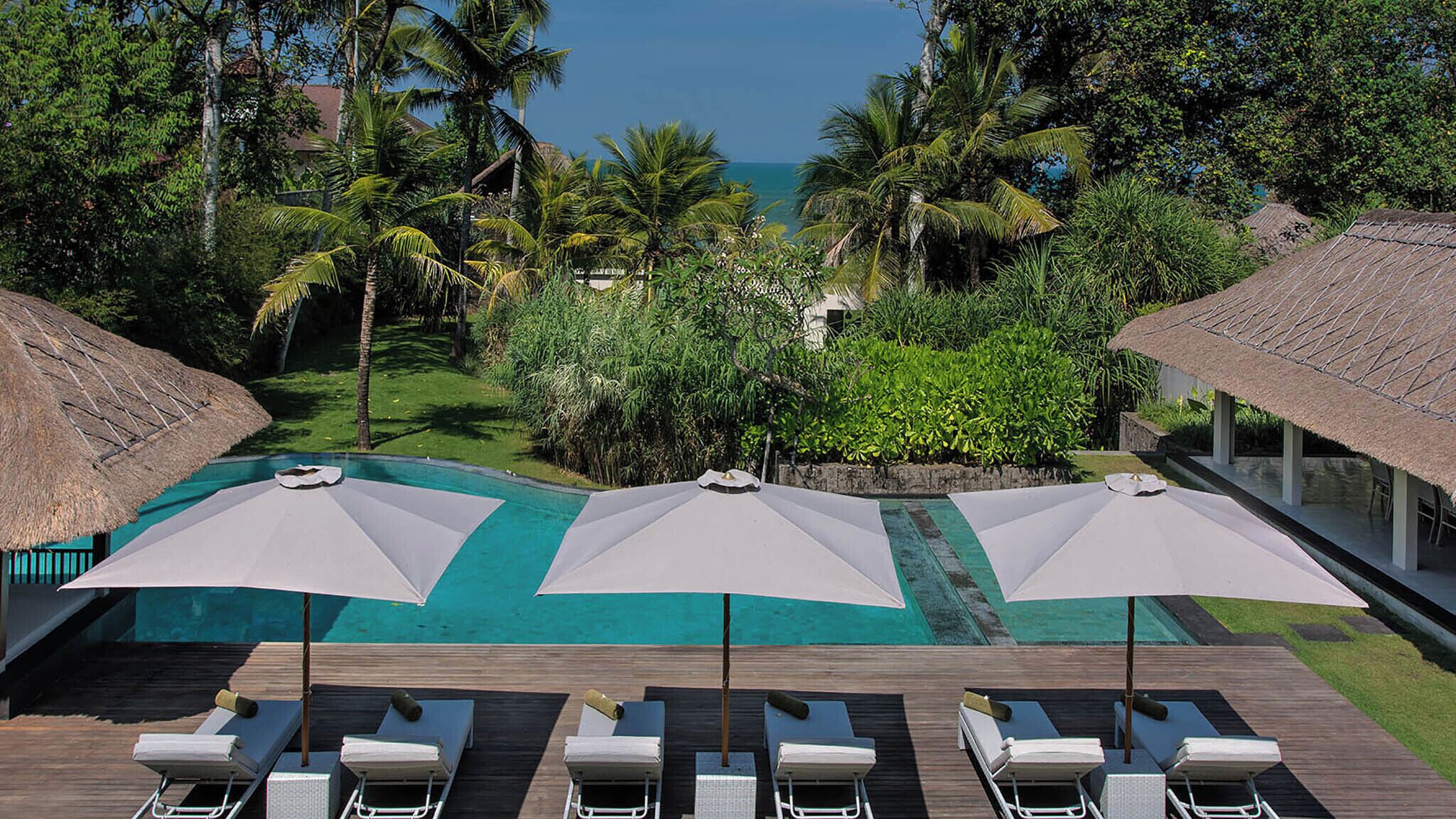 Un espace aménagé au bord de la piscine, avec des chaises longues et des parasols, entouré d’une végétation luxuriante et offrant une vue sur l’océan.