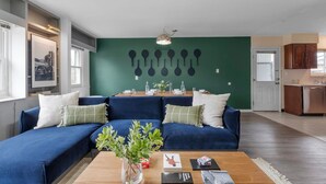 A modern living room with a blue sofa, a wooden coffee table, and a dining area with a green wall and wall-mounted lights. - Walk to OSU • 4BR Ranch • 3 Kings • Pet-Friendly (Columbus)