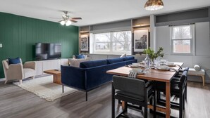A modern living room with a blue sofa, a dining table with chairs, a television, and a ceiling fan. - Walk to OSU • 4BR Ranch • 3 Kings • Pet-Friendly (Columbus)