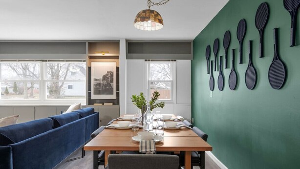A dining area with a wooden table set for four, a blue sofa, and a green wall with tennis rackets. - Walk to OSU • 4BR Ranch • 3 Kings • Pet-Friendly (Columbus)