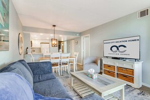 A modern living room with a grey sofa, a wooden coffee table, and a TV on a white cabinet. There is a dining area with a white table and chairs, and a kitchen in the background. - Sea Watch 318 (Ocean City)