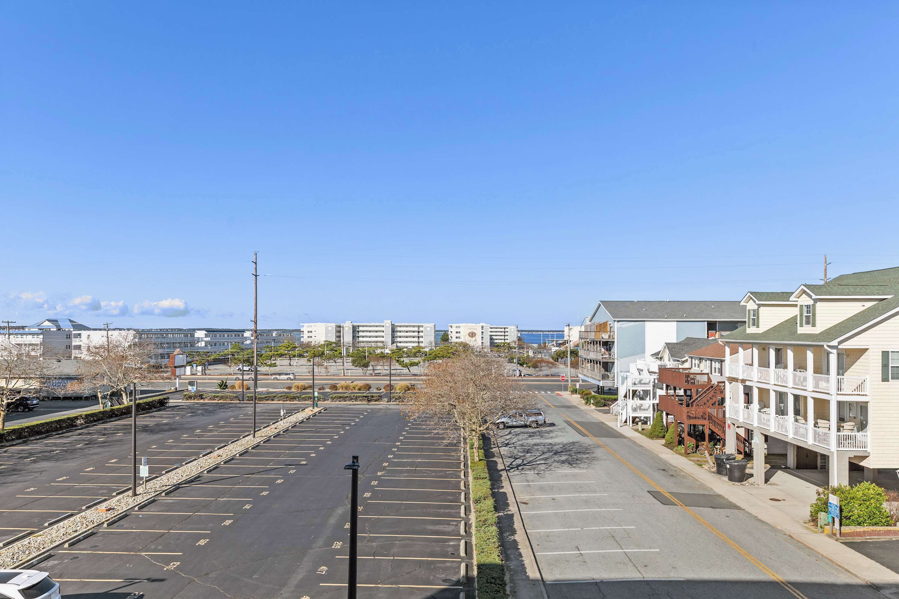 A parking lot with a row of buildings on the right, including a hotel with a balcony and a view of the ocean.