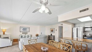 Una cocina moderna con armarios blancos y electrodomésticos de acero inoxidable, un comedor con mesa de madera y sillas de mimbre, y una sala de estar con sofá gris y sillón blanco.