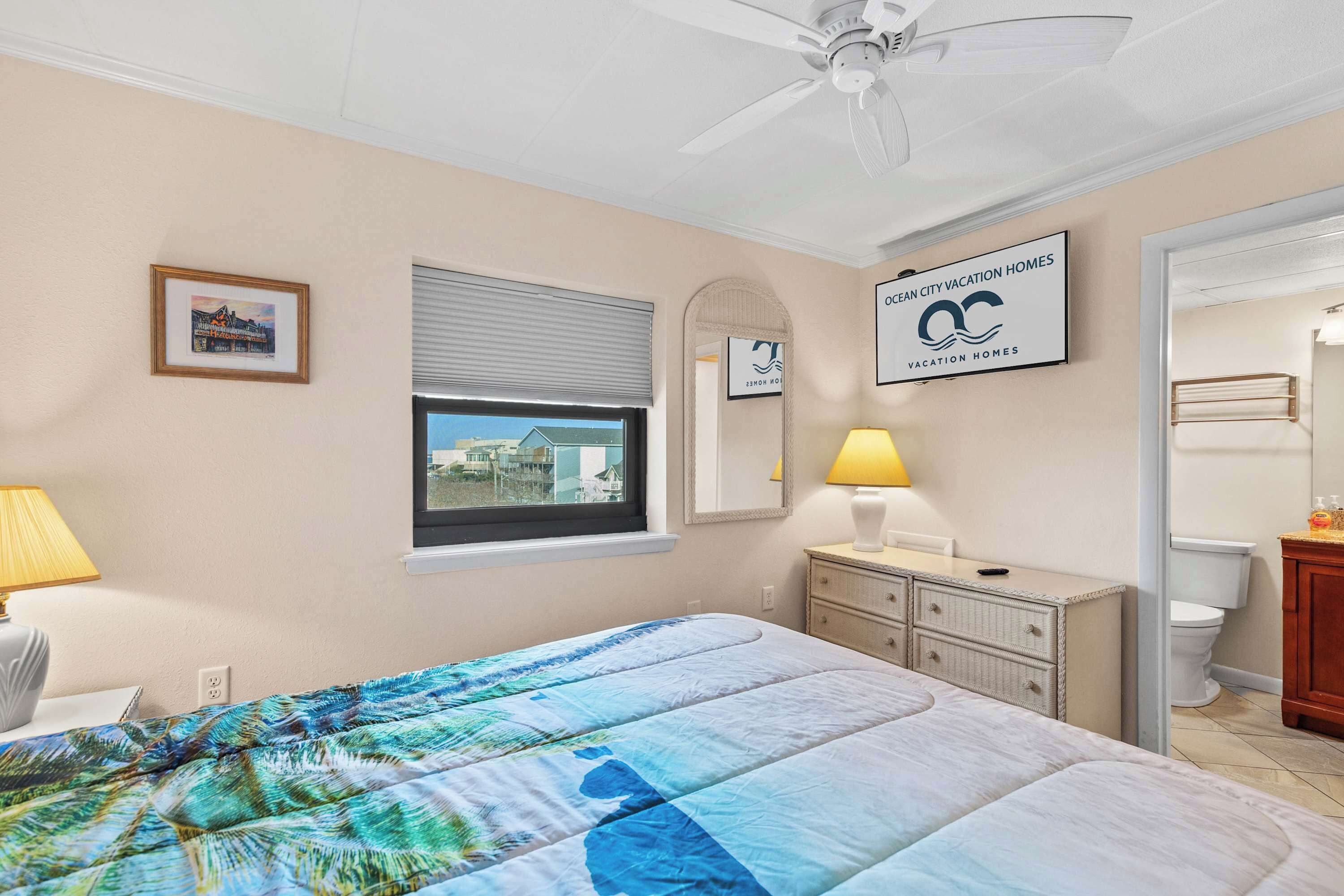 A bedroom with a bed, a window with blinds, a bedside lamp, a dresser, and a framed picture on the wall.