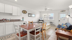A modern kitchen with white cabinets, a dining table with chairs, a ceiling fan, and a living area with a sofa and a TV. - Harbor Breeze #68 (Ocean City)