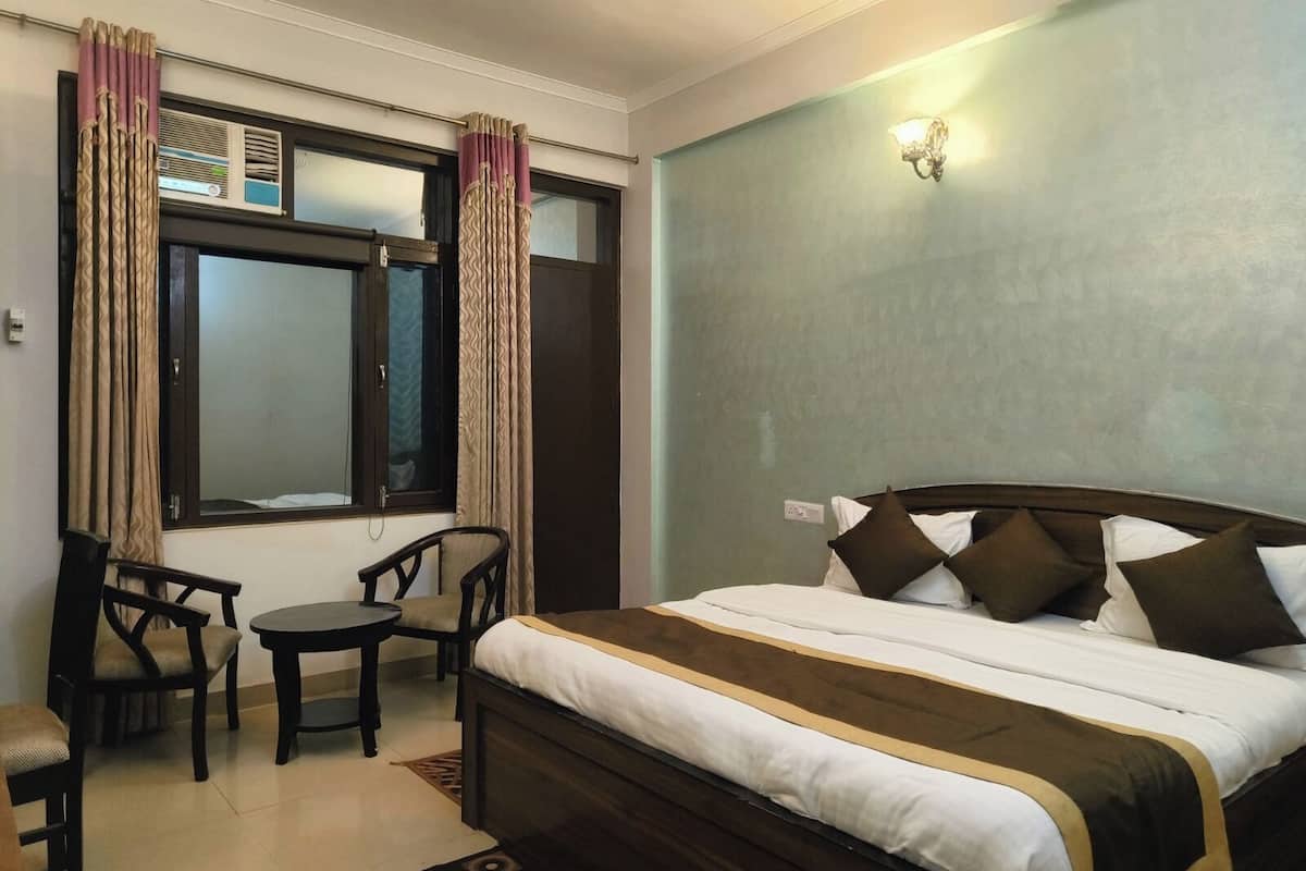 Quarto de hotel com cama, uma pequena mesa, duas cadeiras, janela com cortinas e luminária fixada na parede.
