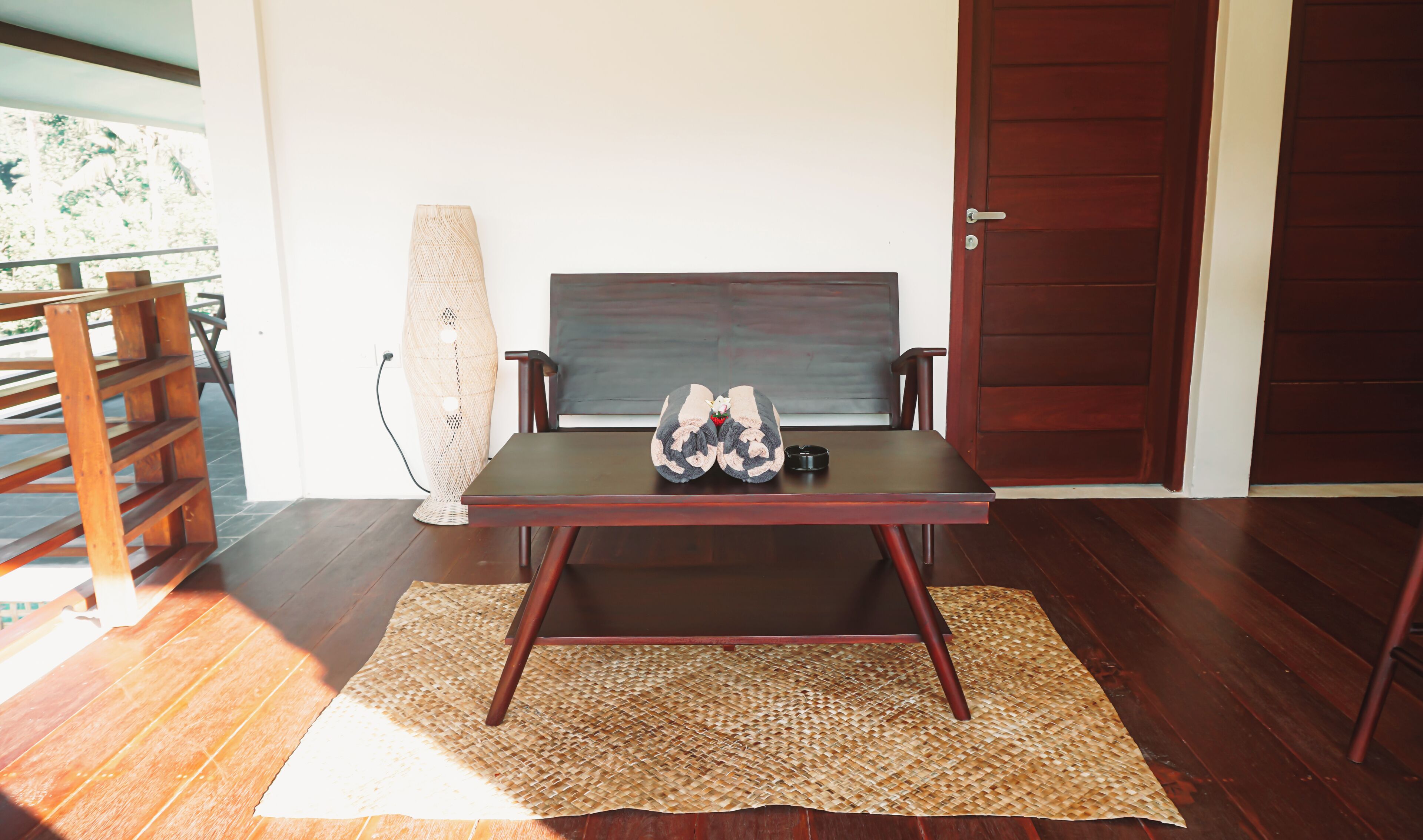 Una habitación con piso de madera, una mesa oscura con un tapete tejido, una silla, una lámpara alta de fibras naturales y una puerta de madera.