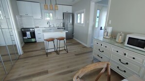 Eine moderne Küche mit weißen Schränken, einem Edelstahl-Kühlschrank und einem eingebauten Backofen. An der Kochinsel stehen zwei Hocker und im Vordergrund ein Holzstuhl.