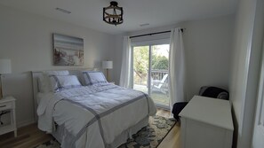 A bedroom with a large bed, a nightstand, a lamp, a painting, a window with curtains, and a view of the outdoors.