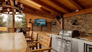 Una terraza de madera con una mesa y sillas, un muro de piedra, una parrilla y una zona de cocina con estufa y fregadero.