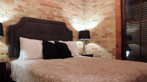 Ein ordentlich bezogenes Bett mit einem gemusterten Bettüberwurf und mehreren Kissen, ein Nachttisch mit Lampe sowie eine Backsteinwand im Hintergrund.