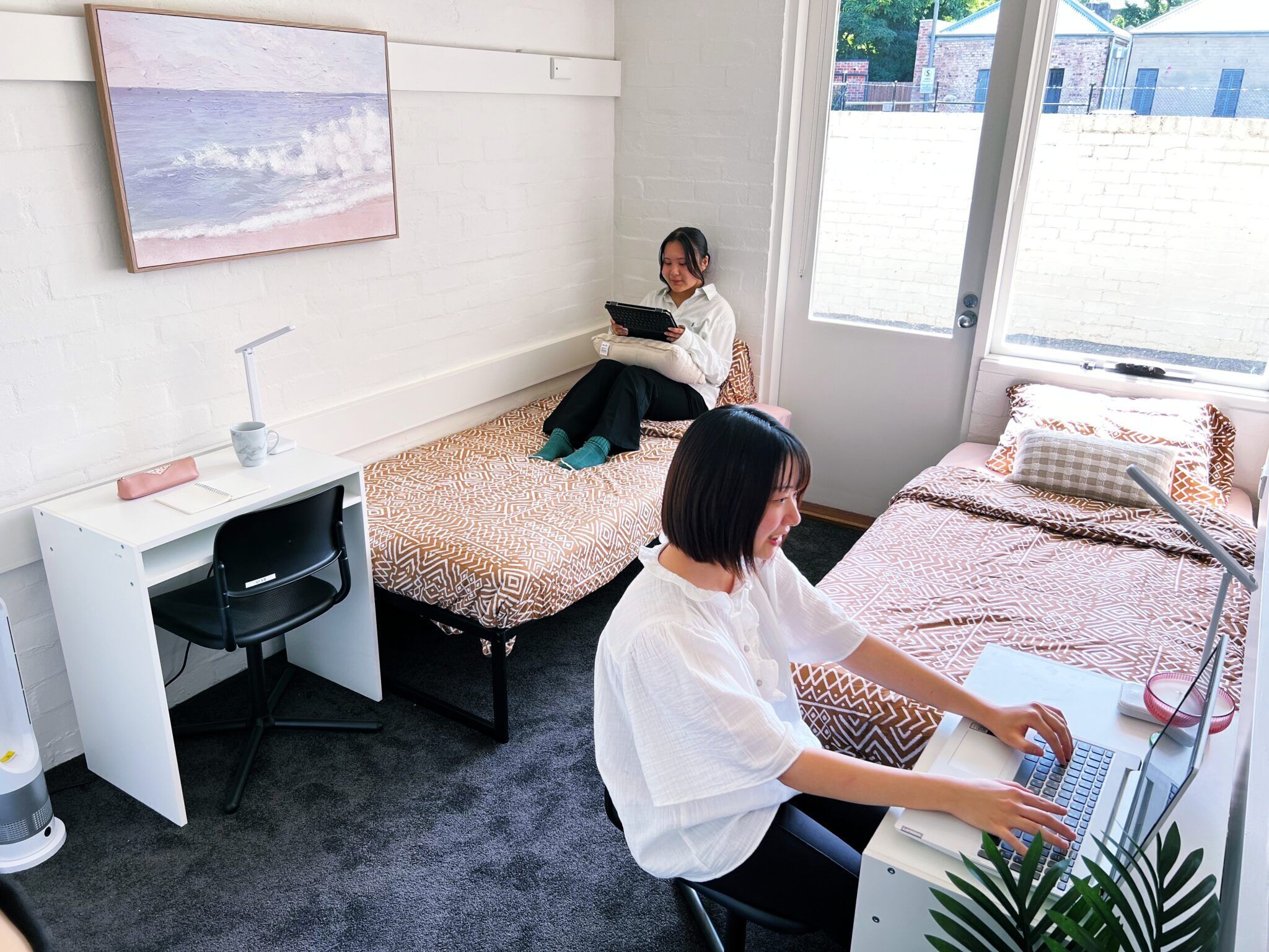 A dormitory room with two single beds, a desk with a chair, a laptop, and a window with a view of buildings.