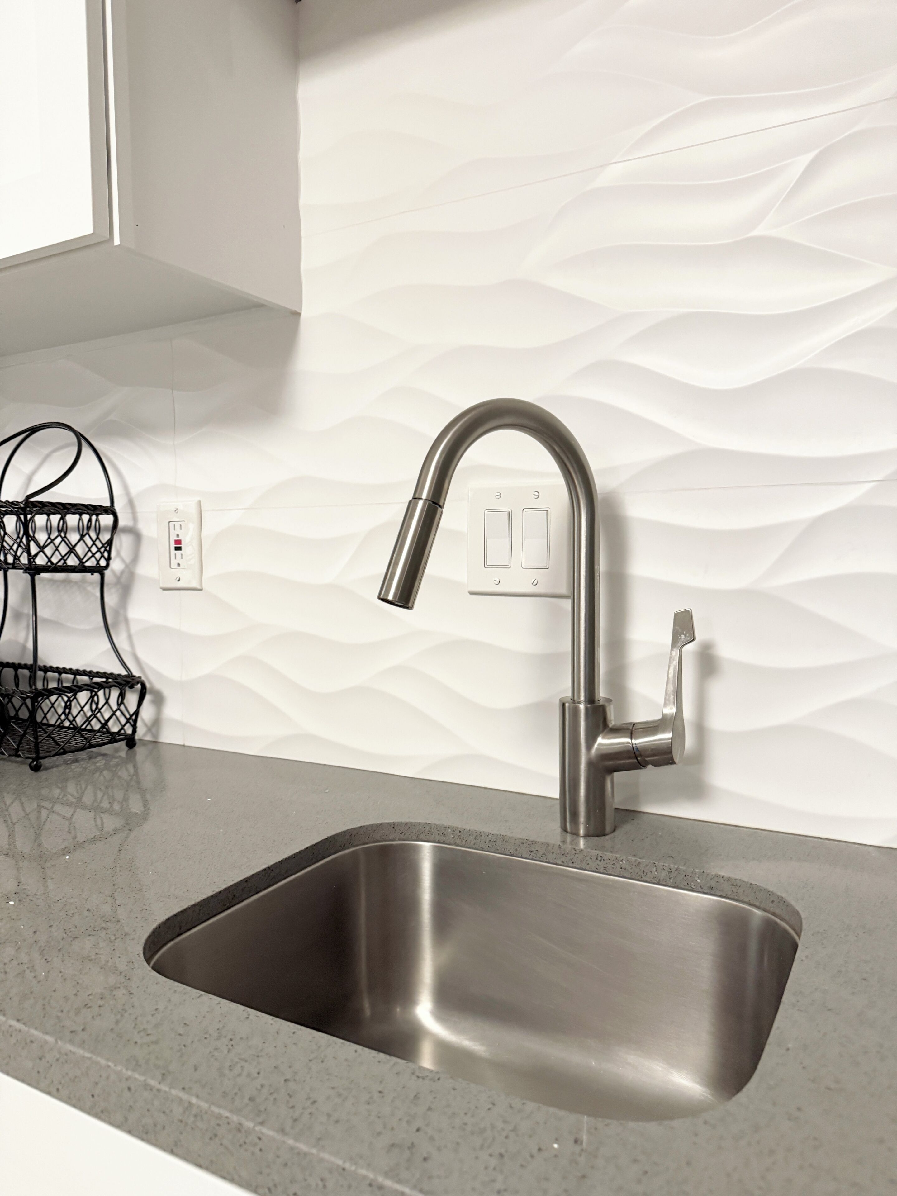 A modern kitchen with a stainless steel sink, a black metal rack, and a white textured backsplash.