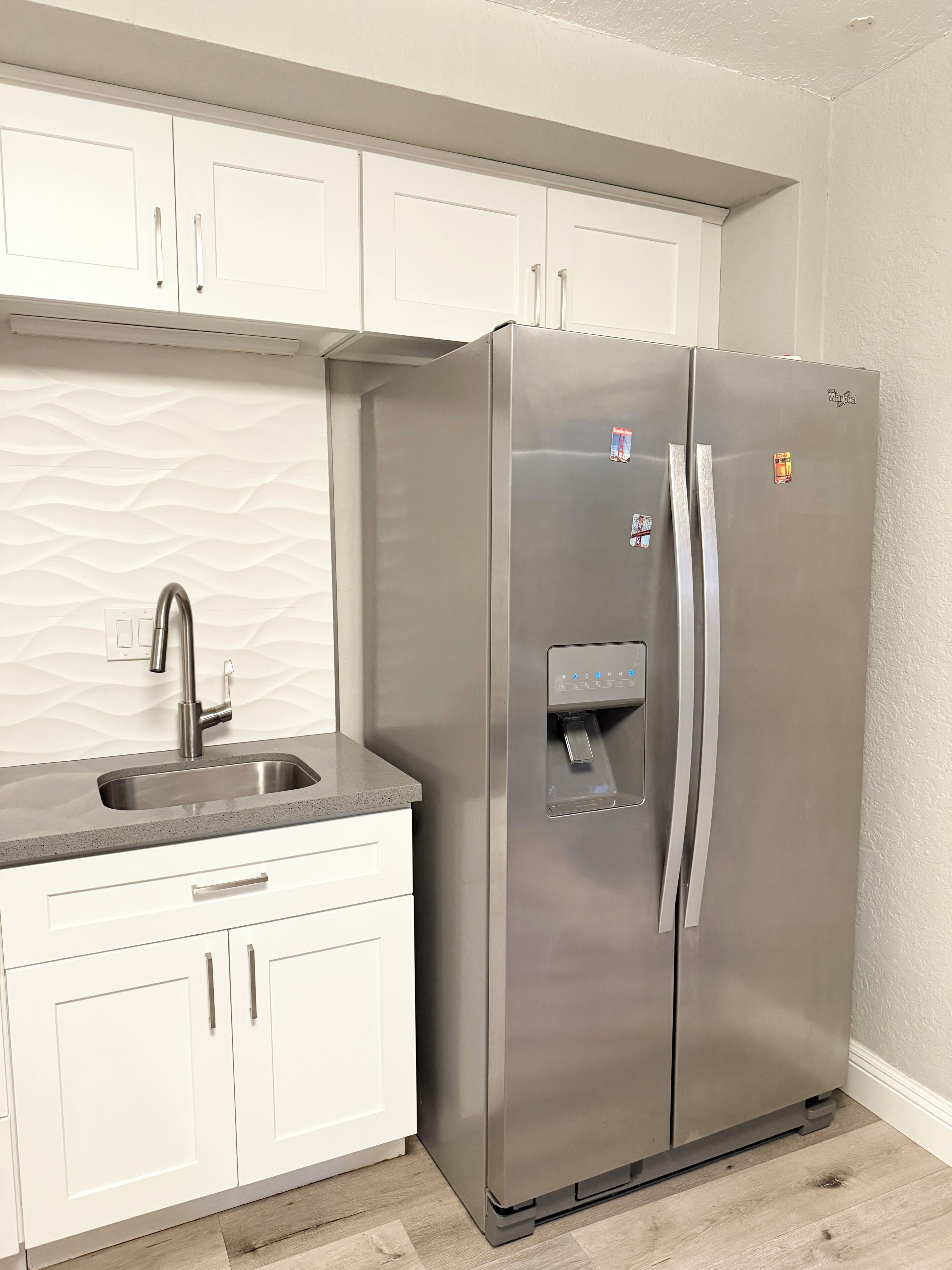 Un réfrigérateur en acier inoxydable doté d’un distributeur d’eau, un évier avec un robinet moderne et des meubles de cuisine blancs.