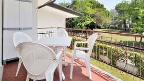 Ein Balkon mit weißem Tisch und Stühlen, einem Metallgeländer und Blick auf einen Park mit Bäumen und einem Zaun.