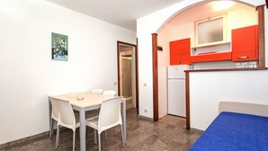 Une pièce compacte comprenant une table et des chaises, un coin cuisine avec des placards rouges et un petit lit doté d’un matelas bleu.