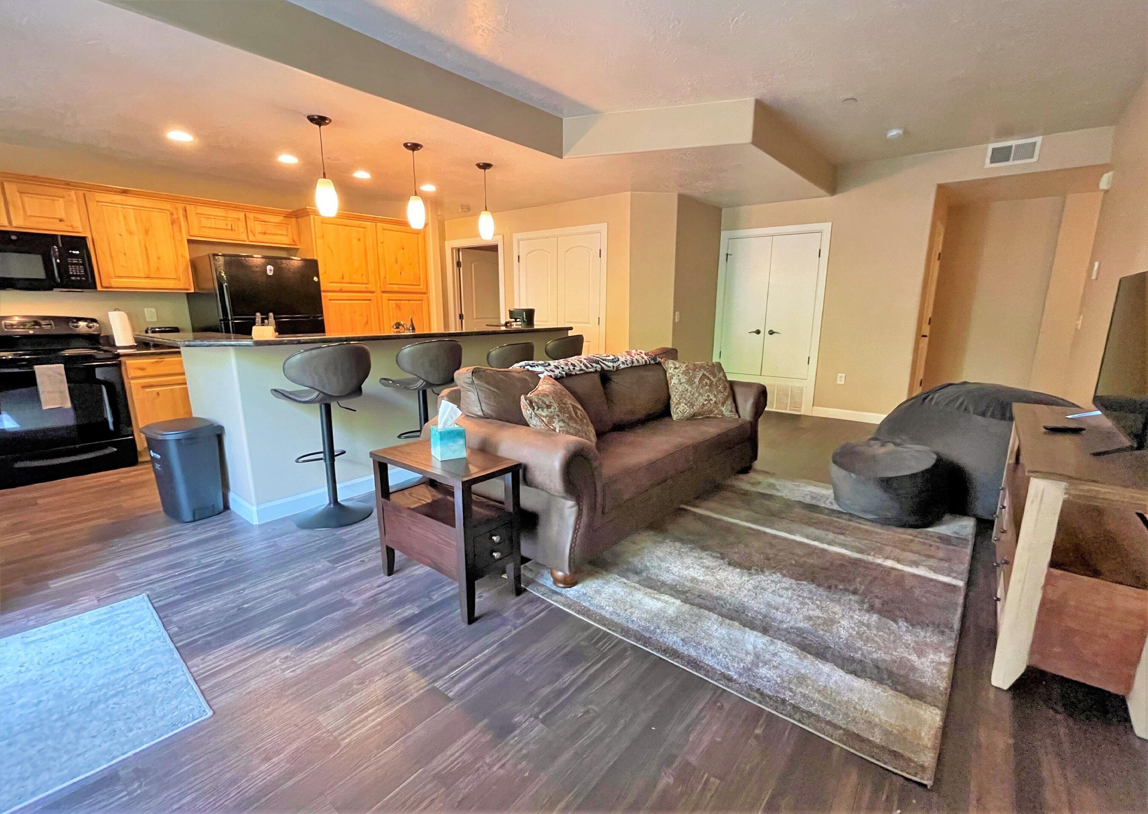 A modern living room with a brown leather sofa, a coffee table, and a rug. There is a kitchen area with wooden cabinets and a refrigerator.