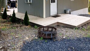 A small, single-story shed with a door and windows, a covered deck area, and a fire pit in the foreground. - Lost River Tiny House, dog-friendly + hot tub (Mathias)