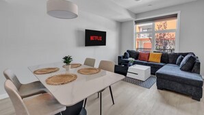 A modern living room with a white dining table, beige chairs, a grey sofa with colorful cushions, a black TV on the wall, and a large window with a view of a balcony. - Modern 2BR Townhome in Belmont with AC & Parking (Calgary)