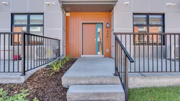 A modern building with a wooden front door, a small porch, and a metal railing. - Modern 2BR Townhome in Belmont with AC & Parking (Calgary)