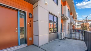 A modern building with a large garage door, a visible apartment number, and a small air conditioning unit on the sidewalk. - Modern 2BR Townhome in Belmont with AC & Parking (Calgary)