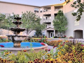 Un edificio de varios pisos con un patio central que presenta una fuente escalonada rodeada de coloridos macizos de flores.