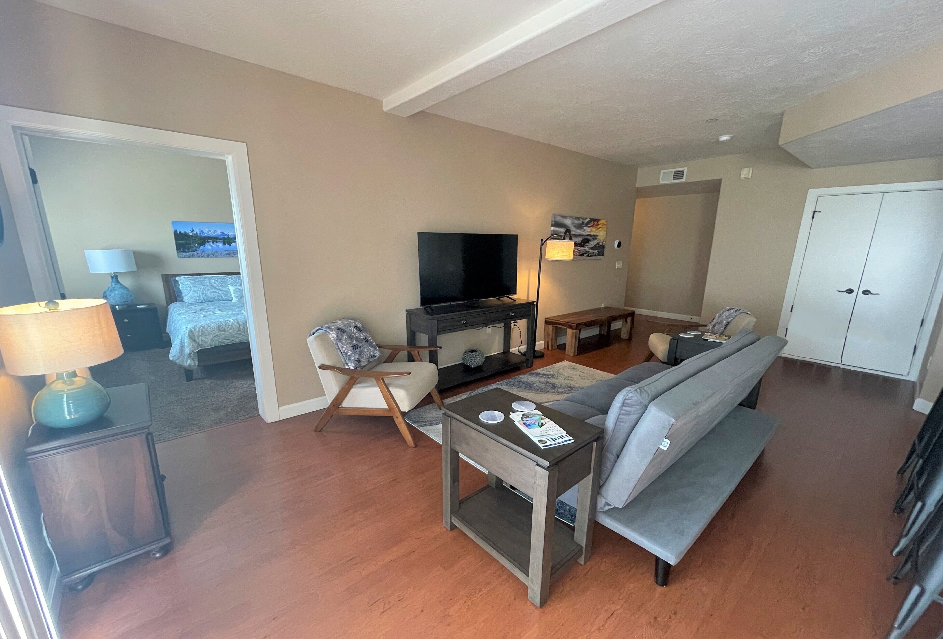 A modern living room with a grey sofa, a TV, a small table with a lamp, and a white wardrobe.