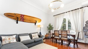 A living room with a grey sofa, a wooden surfboard, a dining table with chairs, and a large window with curtains. - GuestReady - Charming Porto Retreat for Families (Porto)