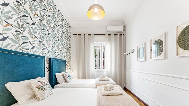 A hotel room with two beds, a blue headboard, a patterned wall, a hanging light fixture, and a view through a window. - GuestReady - Charming Porto Retreat for Families (Porto)