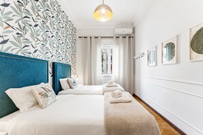 Ein Hotelzimmer mit zwei Betten, einem blauen Kopfteil, einer Wand mit Muster und einer Hängelampe, mit Blick durch ein Fenster.