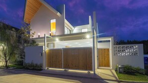 A modern two-story house with a large wooden door, a balcony, and a well-lit entrance. - Luxpride4  (Si Sunthon)