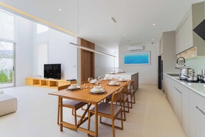 A modern kitchen with a dining area, featuring a long wooden table set for six, a built-in oven, and a large window providing a view of the outdoors. - Luxpride4  (Si Sunthon)
