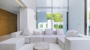 A modern living room with a sectional sofa, a round coffee table, and large windows offering a view of greenery outside. - Luxpride4  (Si Sunthon)