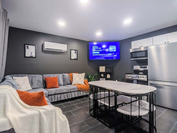 Una sala de estar moderna con un sofá gris, una mesa de comedor y una zona de cocina con nevera y horno.