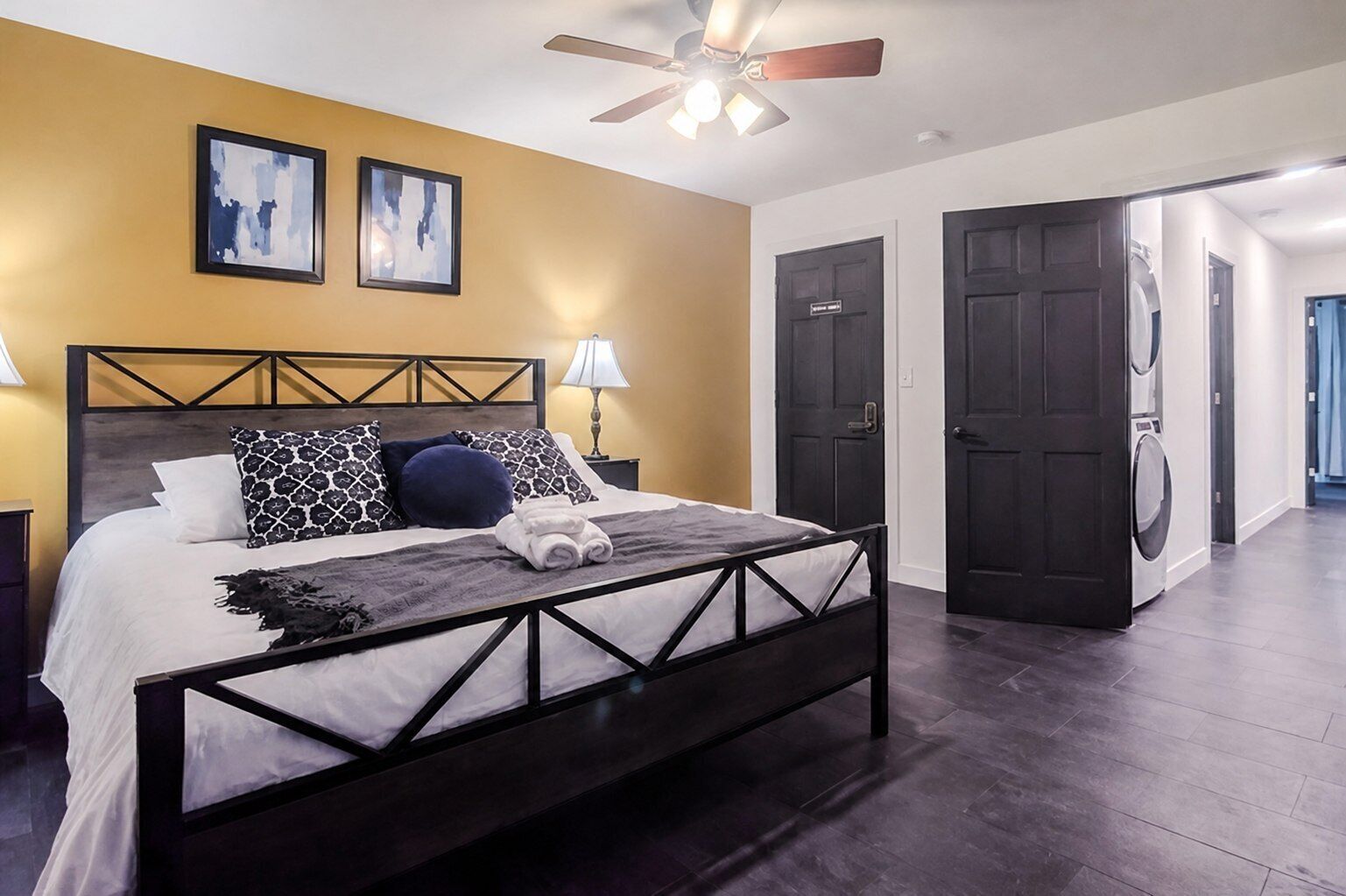 Une chambre à coucher comprenant un lit, un ventilateur de plafond, deux tableaux encadrés et un coin buanderie avec un lave-linge.