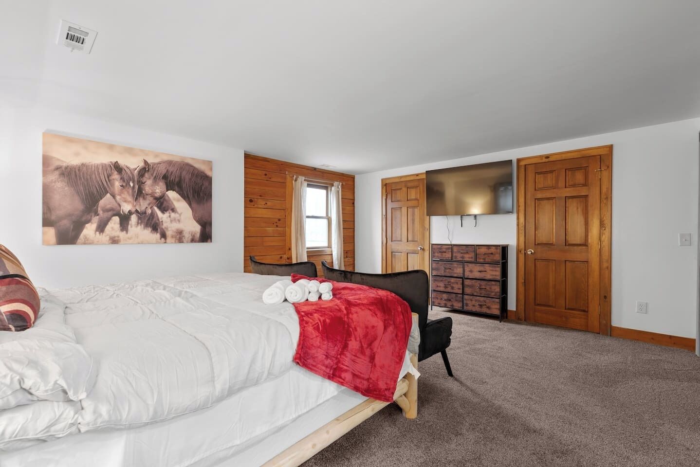 A bedroom with a bed, a red blanket, a wooden dresser, a TV, and a painting of horses.