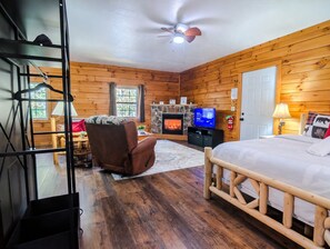 A wooden cabin interior with a bed, a television, a fireplace, and a ceiling fan. - Couples Cabin with Hot Tub, Grill, Firepit & EV (Logan)