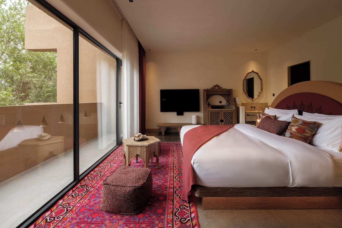 A modern hotel room with a large bed, a small table, a TV, and a balcony with a view of trees.