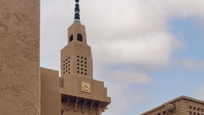 A person walking in an open courtyard with a tall building featuring a minaret in the background.