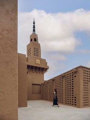 A person walking in an open courtyard with a tall building featuring a minaret in the background.