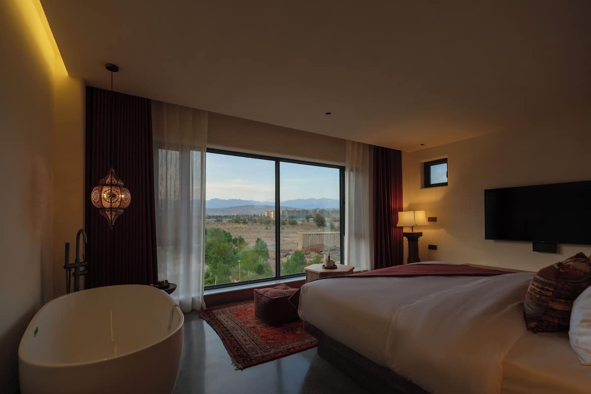 A modern hotel room with a large bed, a bathtub, a flat-screen TV, and a view of the cityscape.