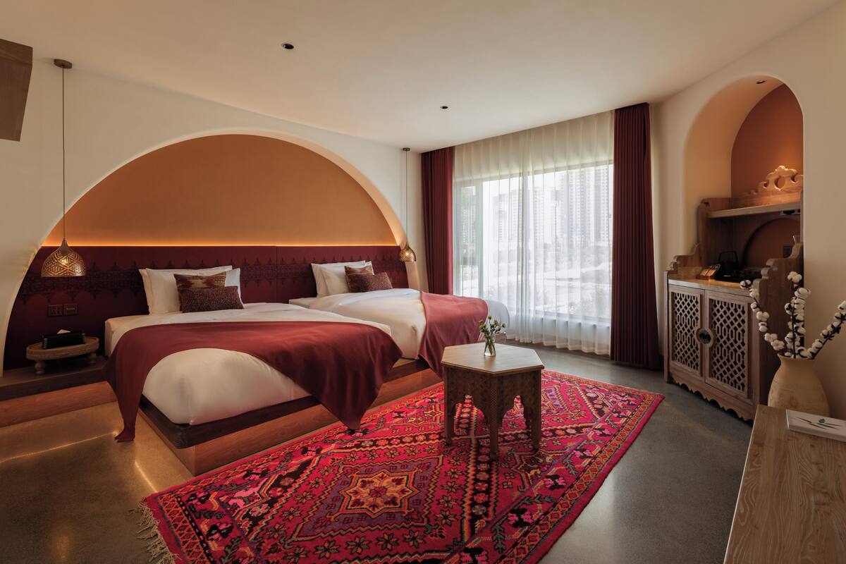 A hotel room with a large bed, a small table, a decorative rug, and a view of the city through large windows.
