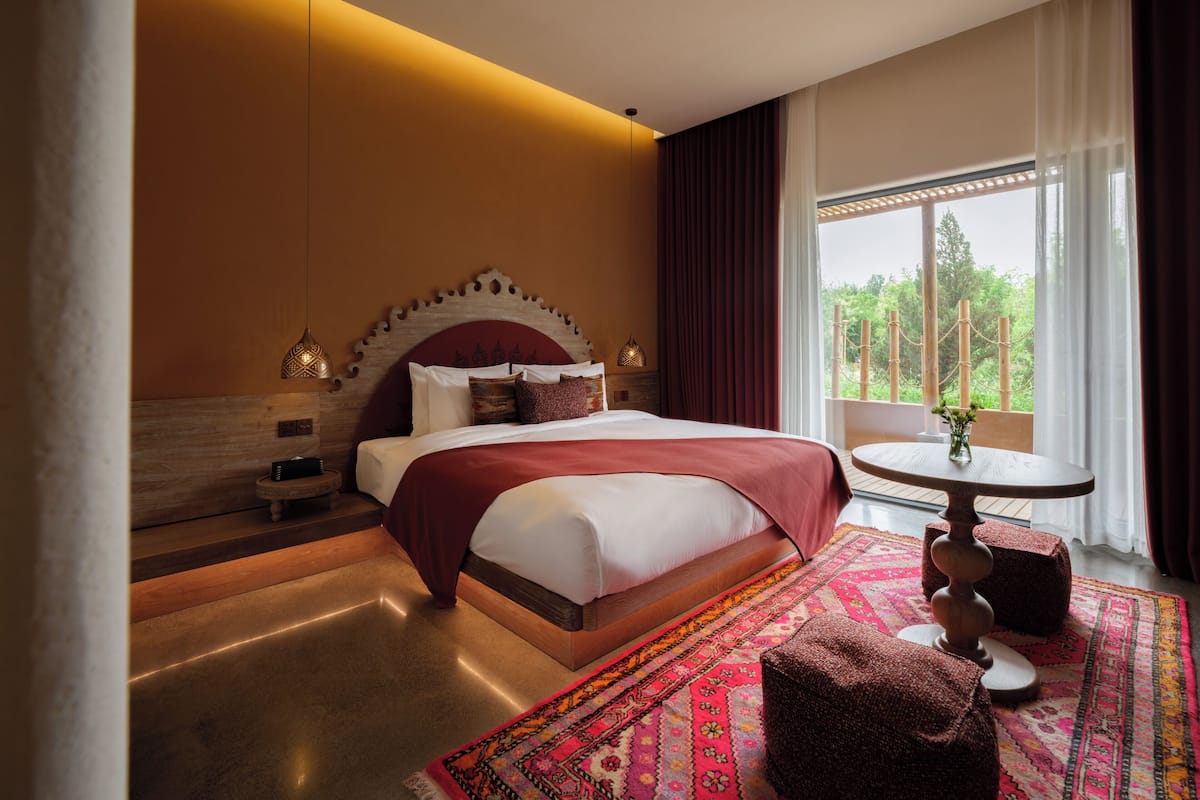 A modern hotel room with a large bed, a round table, and a view of greenery outside.