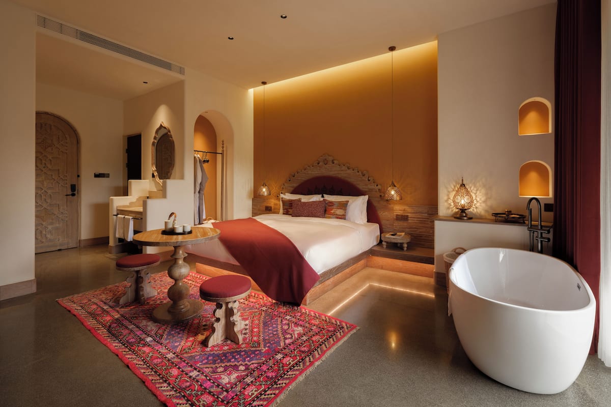 A modern hotel room with a large bed, a sitting area with a table, a large bathtub, and a decorative rug.