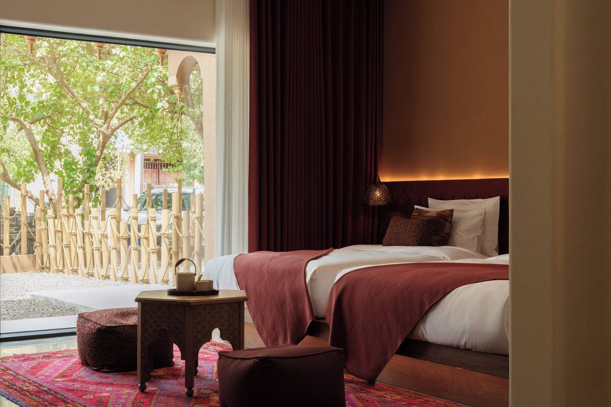 A modern hotel room with a large window, a bed with a red blanket, a small side table, and a colorful rug.