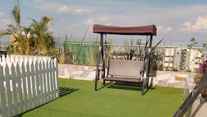 Un espacio patio con césped artificial, un conjunto de columpios, una valla de piquetes blancos y vistas de edificios al fondo.
