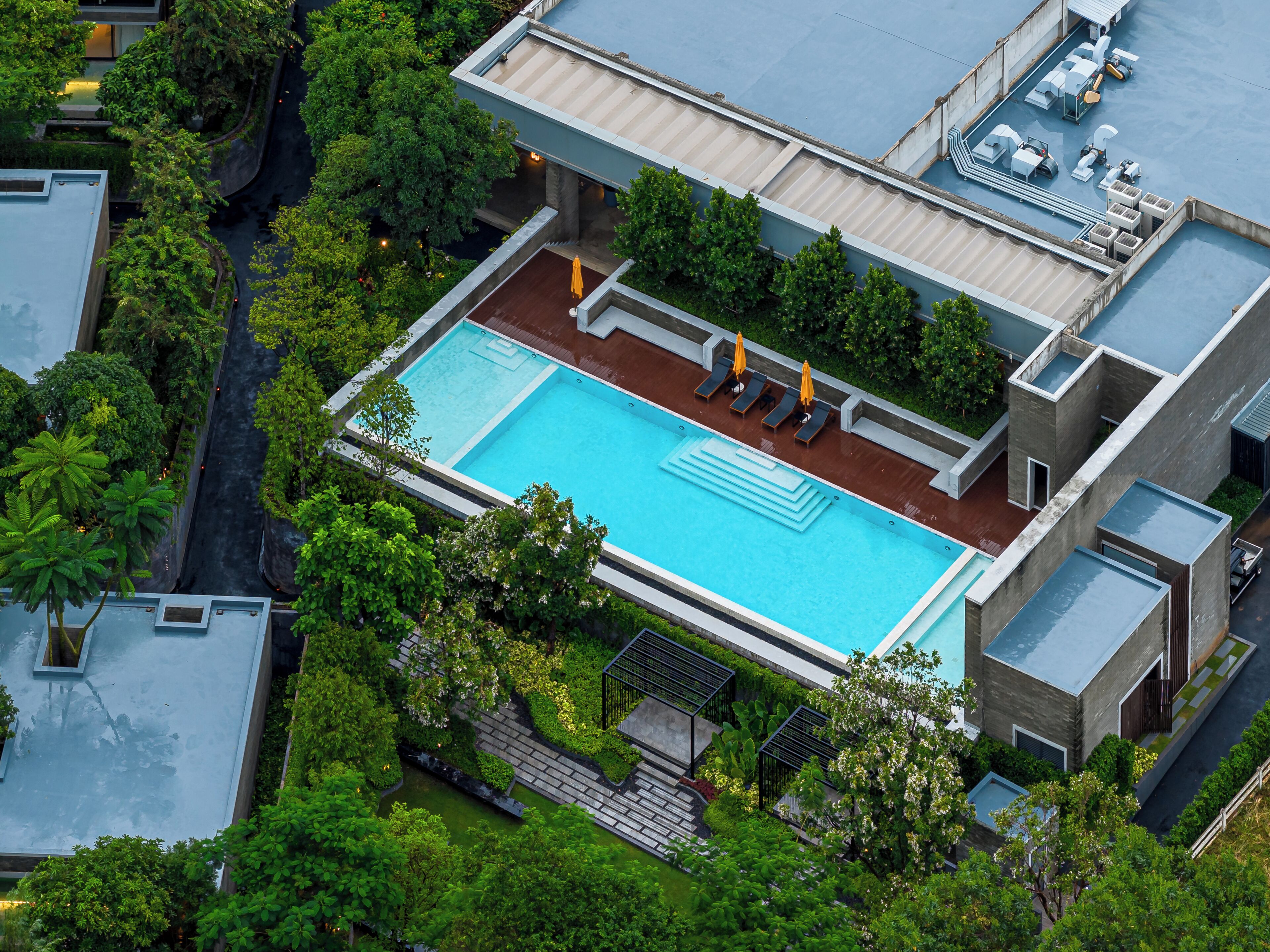 An aerial view of a modern building with a rectangular swimming pool surrounded by lush greenery and a covered outdoor area with lounge chairs and umbrellas.