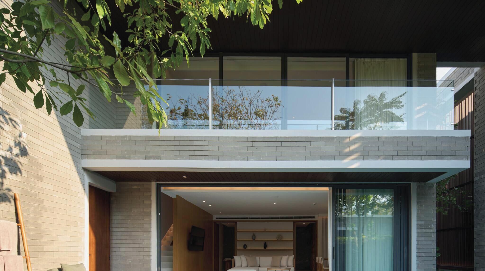 Uma casa moderna de dois andares com varanda de vidro, fachada de tijolos e interior visível com sofá e área de jantar.