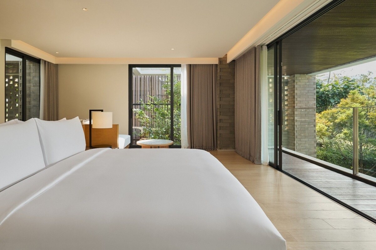 Une chambre d’hôtel moderne avec un grand lit, une table de chevet, une petite table ronde et un balcon donnant sur un paysage verdoyant.